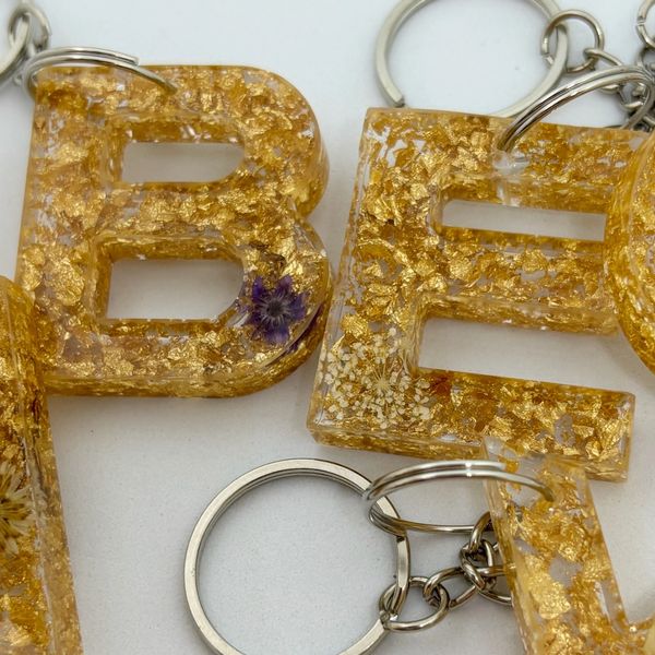 Golden resin keychains shaped as letters with embedded floral details.