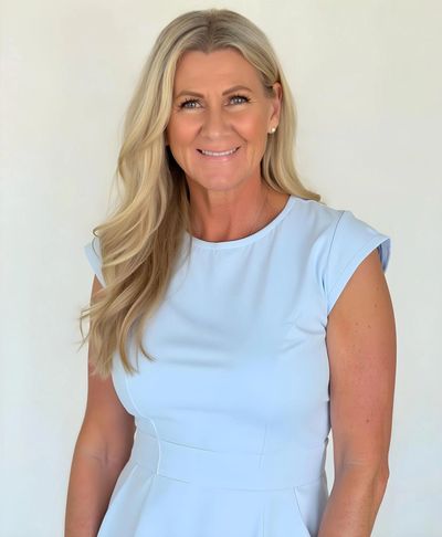 Smiling woman with long blonde hair in a light blue dress against a plain background.