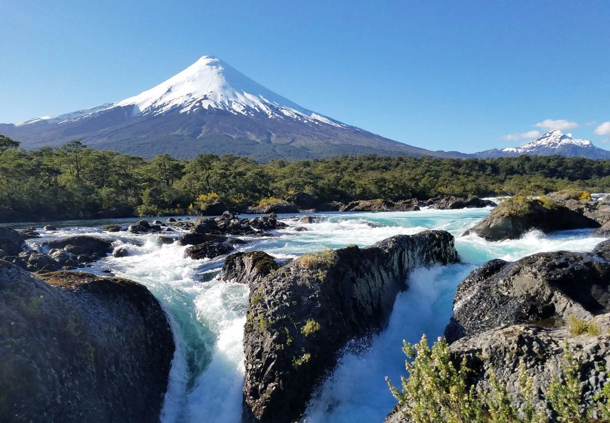 Snow-capped volcano with flowing river and rocks in foreground under clear blue sky.