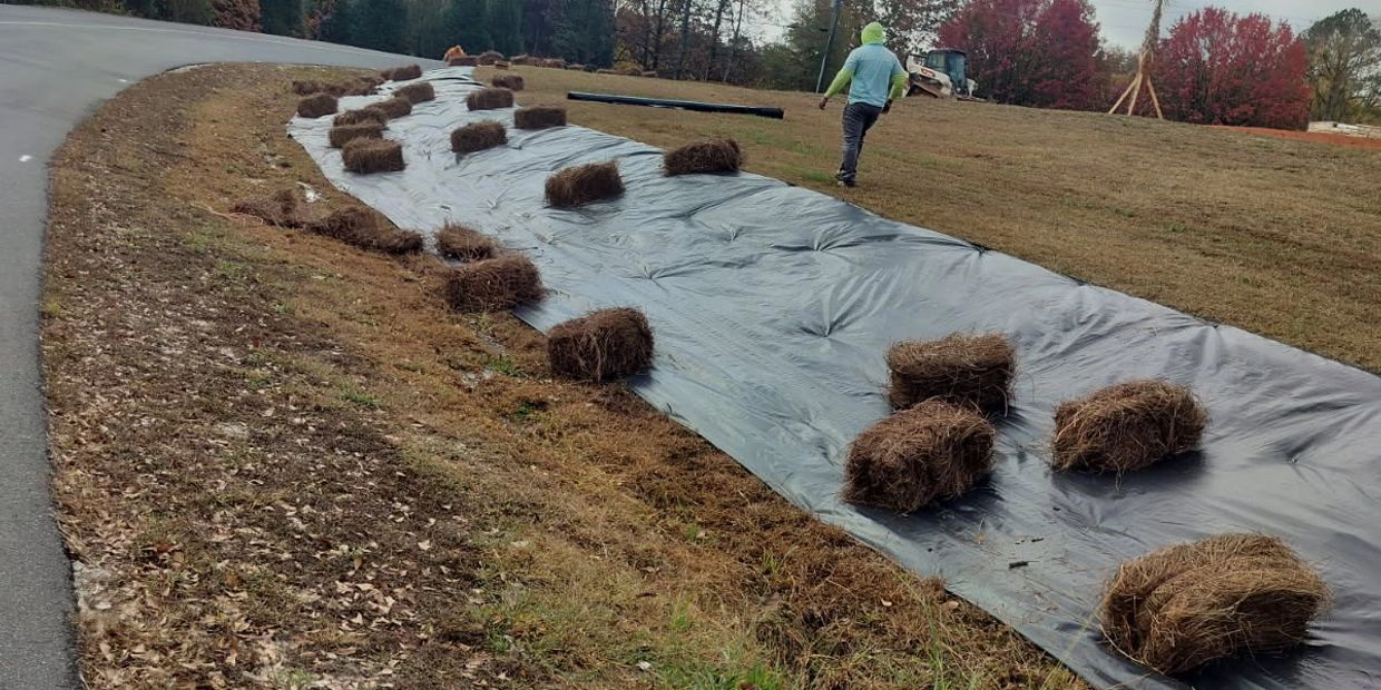 A person walking near a black plastic sheet held down by hay bales on a slope.