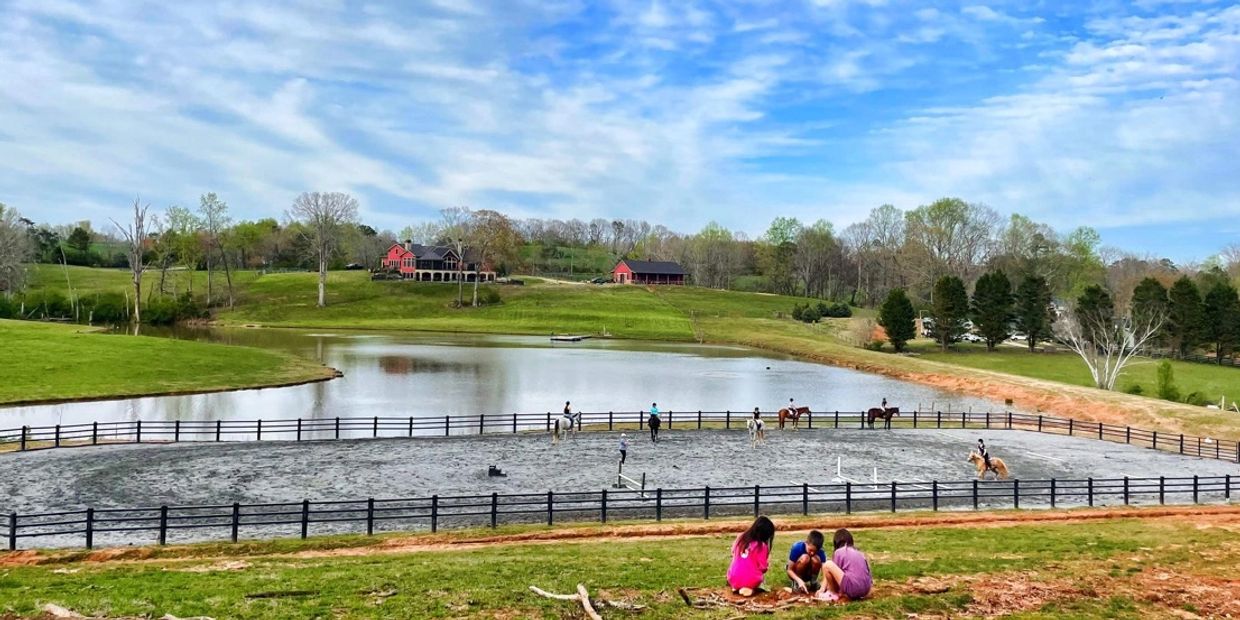 Children playing by a fenced horse riding arena near a lake.