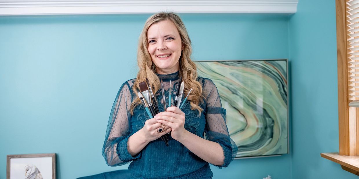 Smiling woman holding paintbrushes in a blue room.