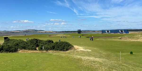 Golfers play on a sunny course with blue skies and distant hills.