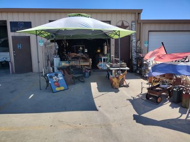 Outdoor market stall with various items under a canopy in front of a garage.