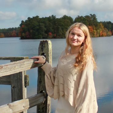 A woman in light clothing stands by a wooden fence near a calm lake surrounded by autumn trees.