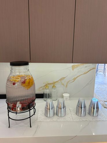 A glass beverage dispenser with fruit and four metal cups on a table.