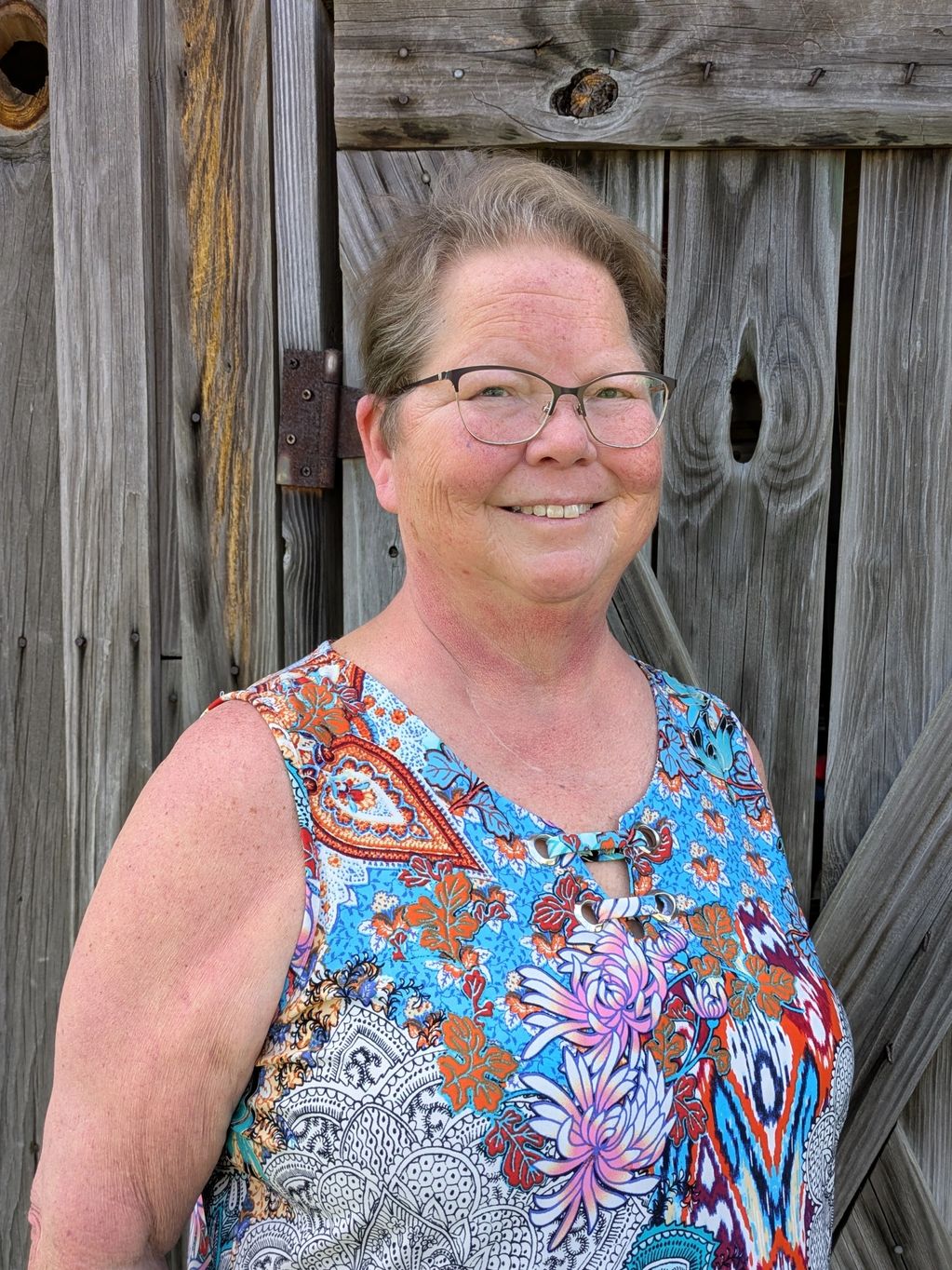 Smiling woman in glasses stands against rustic wooden backdrop.