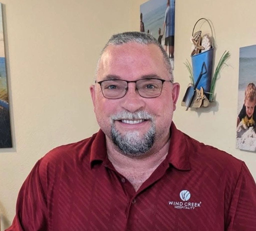 Smiling man wearing glasses and a red Wind Creek Hospitality shirt indoors.