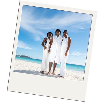 Three women in white dresses posing on a sunny beach with blue sky.
