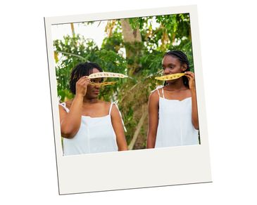 Two women holding long yellow leaves in front of their faces outdoors.