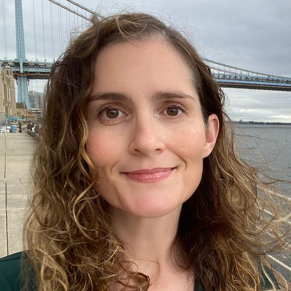 Smiling woman with curly hair standing near a waterfront and bridge.
