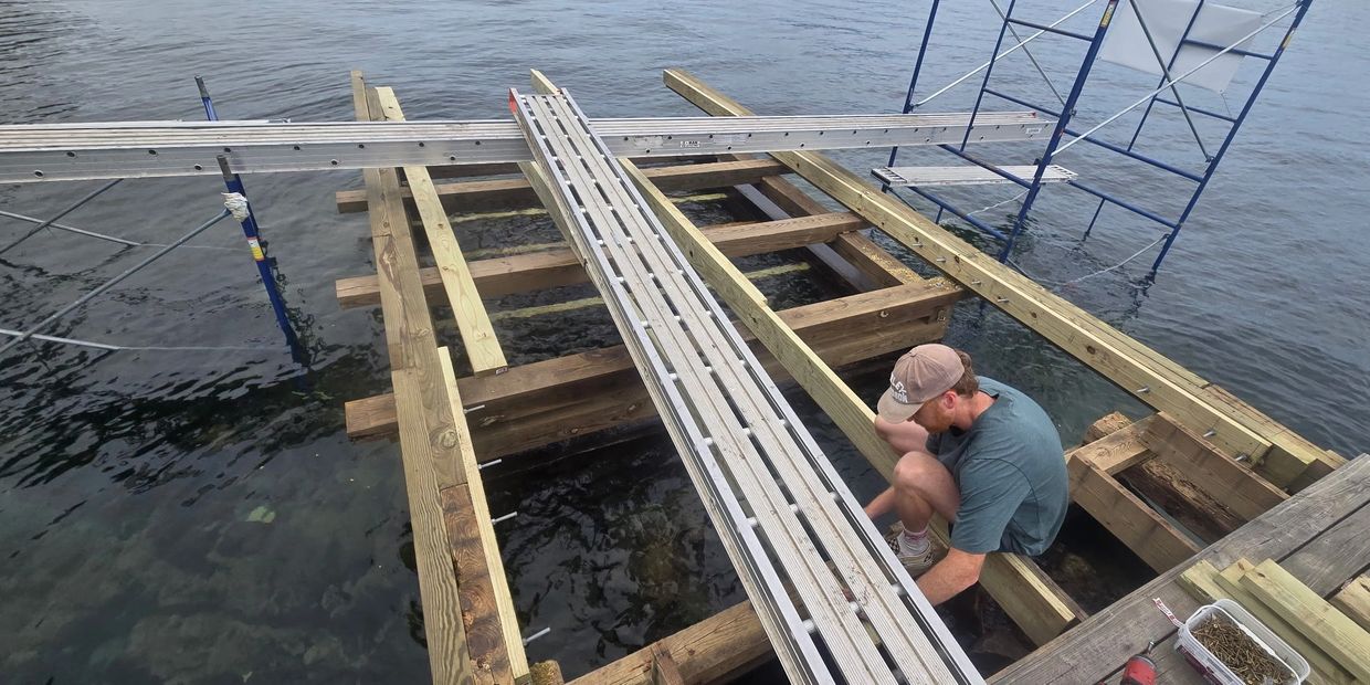 Man building wooden dock over water using scaffolding and planks.