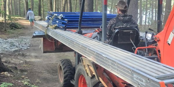 A tractor carrying long metal pipes on a forest path with a person walking ahead.