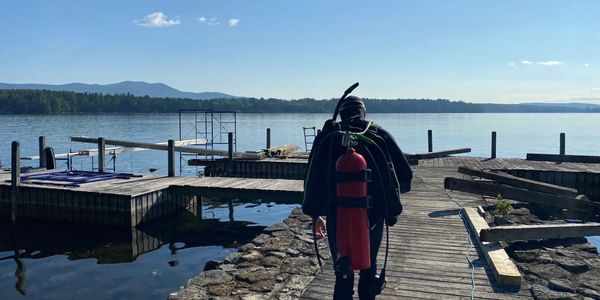 A scuba diver walking on a dock towards a lake with mountains in the background.