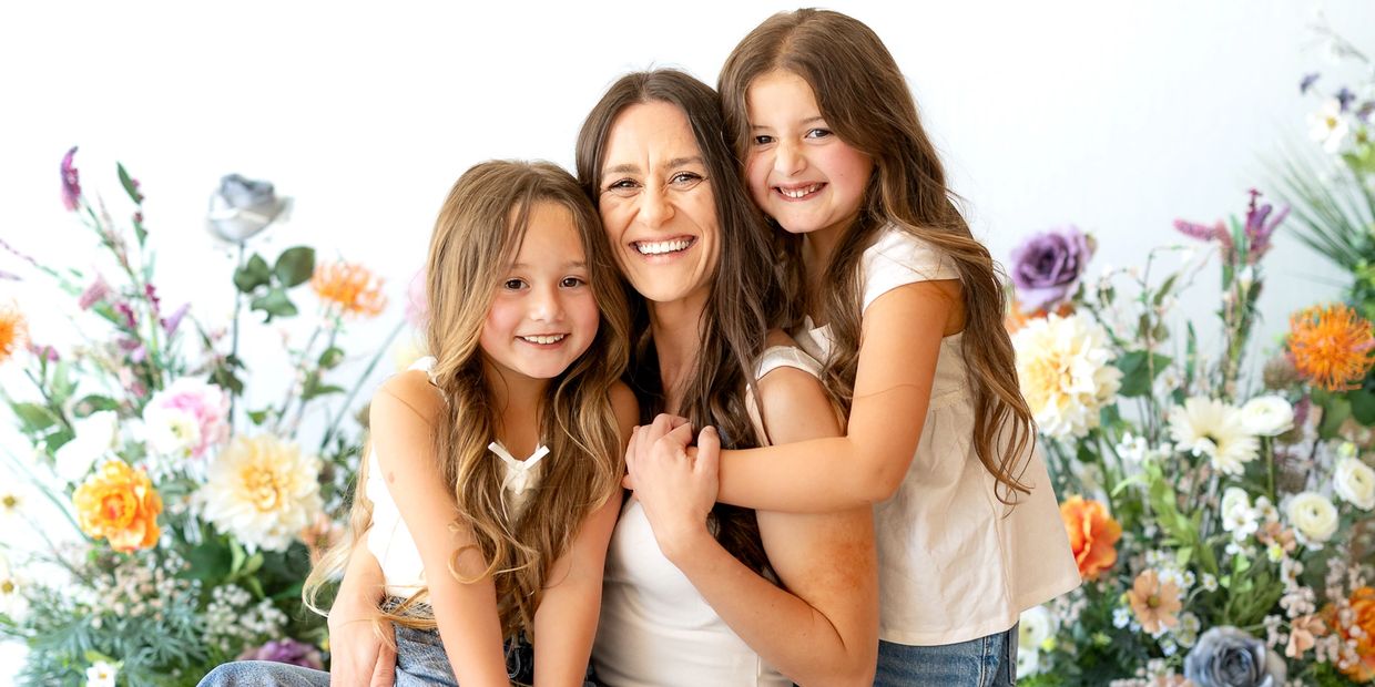 A joyful mother hugs her two smiling daughters surrounded by colorful flowers.