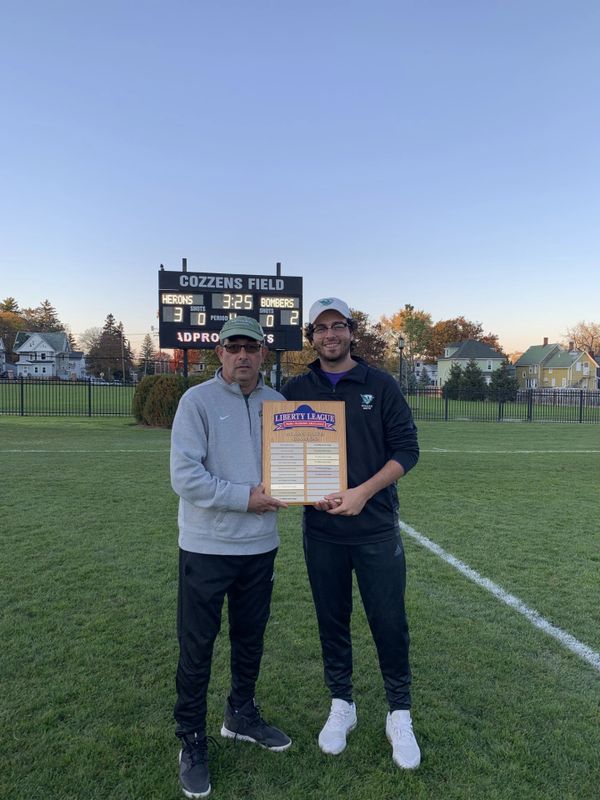Two men hold a Liberty League plaque on a sports field with a scoreboard in the background.