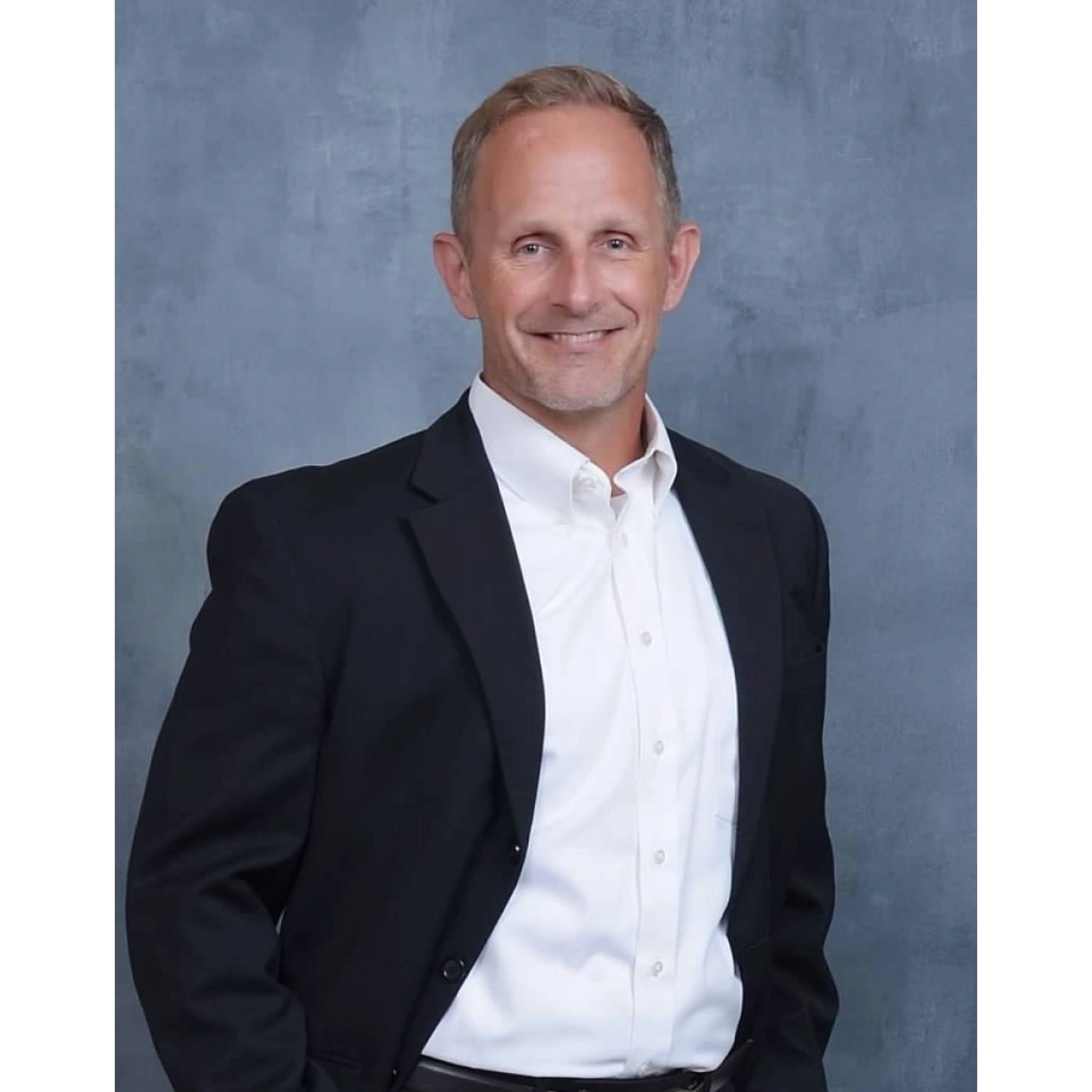 Man in a black suit jacket and white shirt smiling against a gray background.