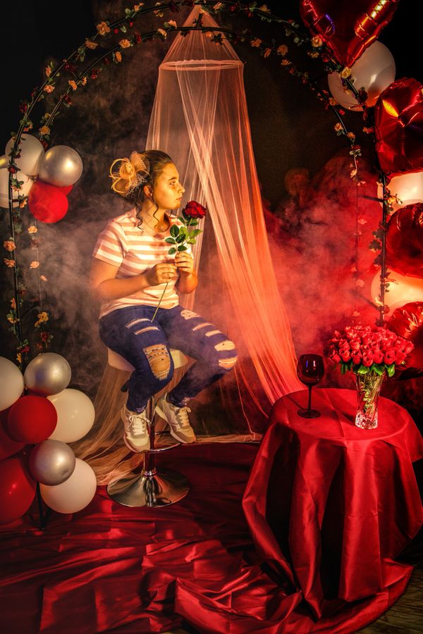 Young woman holding a red rose, sitting in a romantic, decorated setting with balloons and flowers.