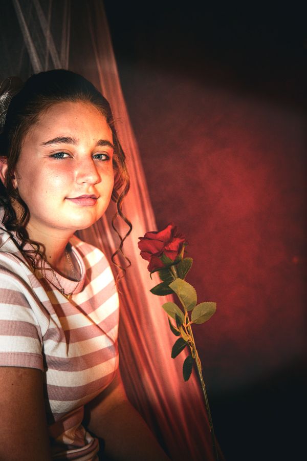 Young girl with curly hair holding a red rose, sitting by a curtain.