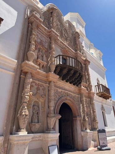 Historic ornate stone entrance with carved details and wooden balconies against a bright blue sky.
