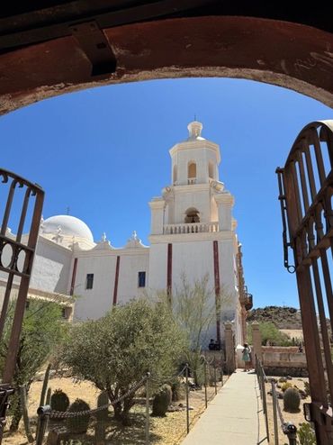 View of a white historic mission church framed by an arched gate under a clear blue sky.