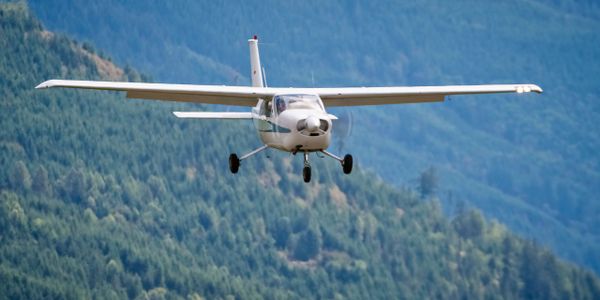 A small white airplane flying over a forested mountainous area.