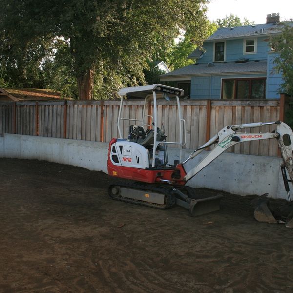 Small excavator parked on a dirt yard near a wooden fence and blue house.