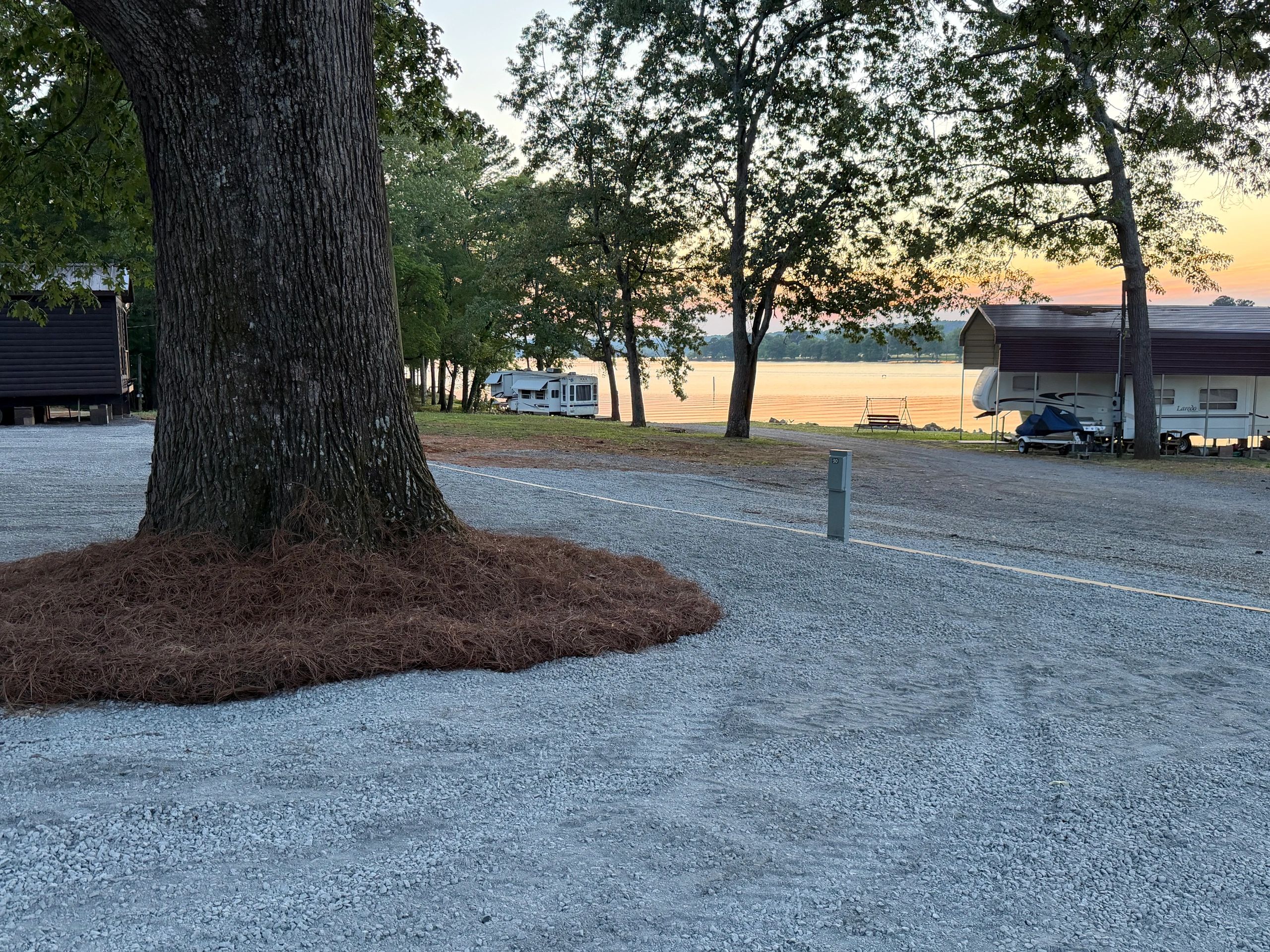 Sunset at a lakeside RV park with trees and gravel paths.