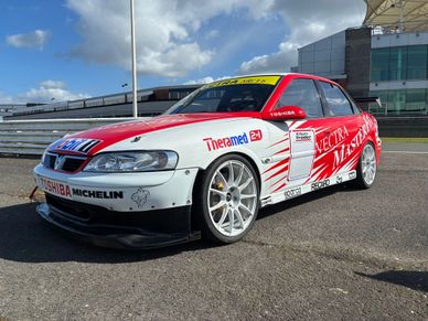 Red and white race car with sponsor logos parked on asphalt under a partly cloudy sky.
