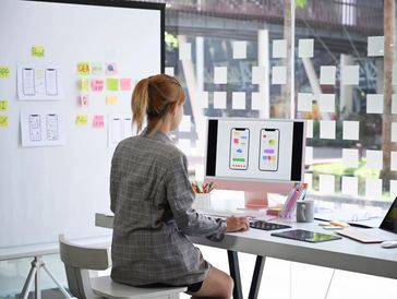 Woman designing mobile app interfaces at her desk with sticky notes on the wall.