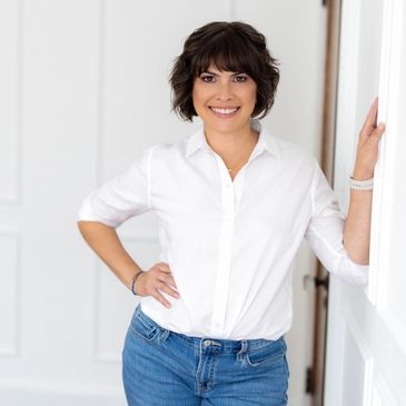 Smiling woman in white shirt and jeans leaning against a wall.