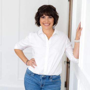 Smiling woman in white shirt and jeans leaning against a wall.
