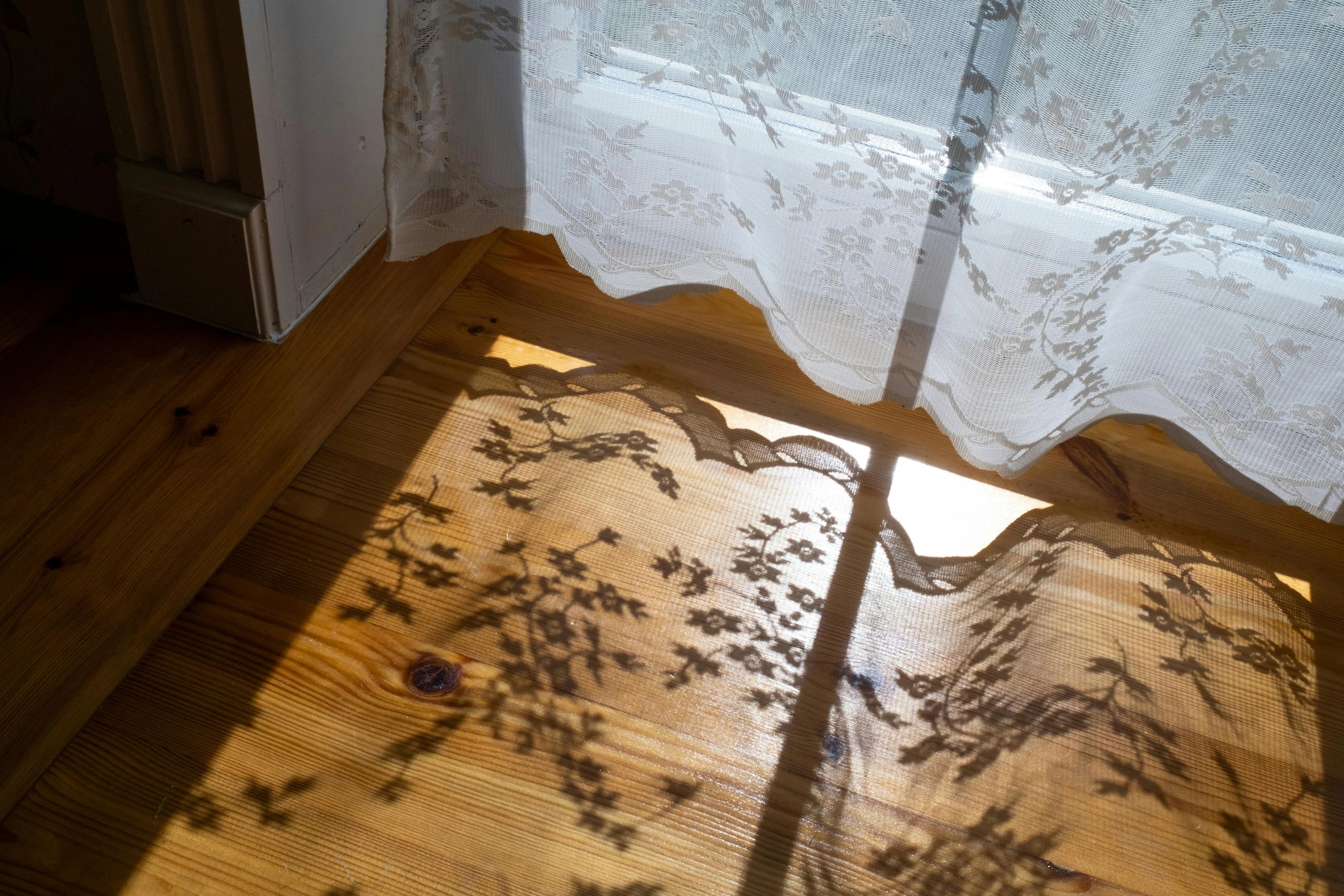 Sunlight casting floral lace curtain shadows on a wooden floor.