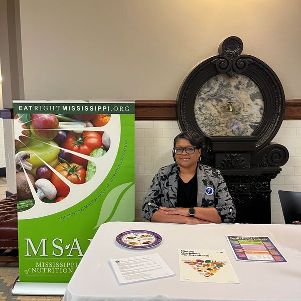 Woman sitting at a nutrition education booth with a colorful fruit and vegetable poster.
