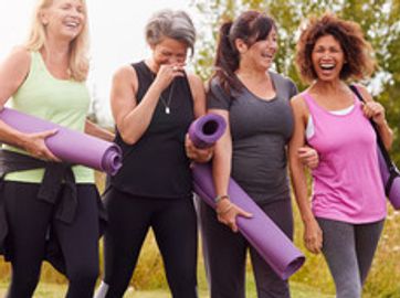 Four women holding yoga mats, laughing together outdoors.