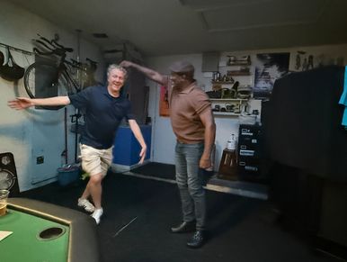 Two men dancing joyfully in a cozy room with a bicycle on the wall.