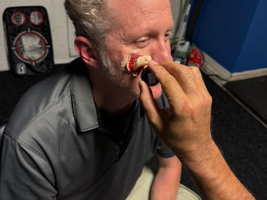 A person applying a realistic fake nose prosthetic to another man's face.