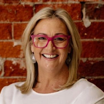Smiling woman with blonde hair and pink glasses against a brick wall.