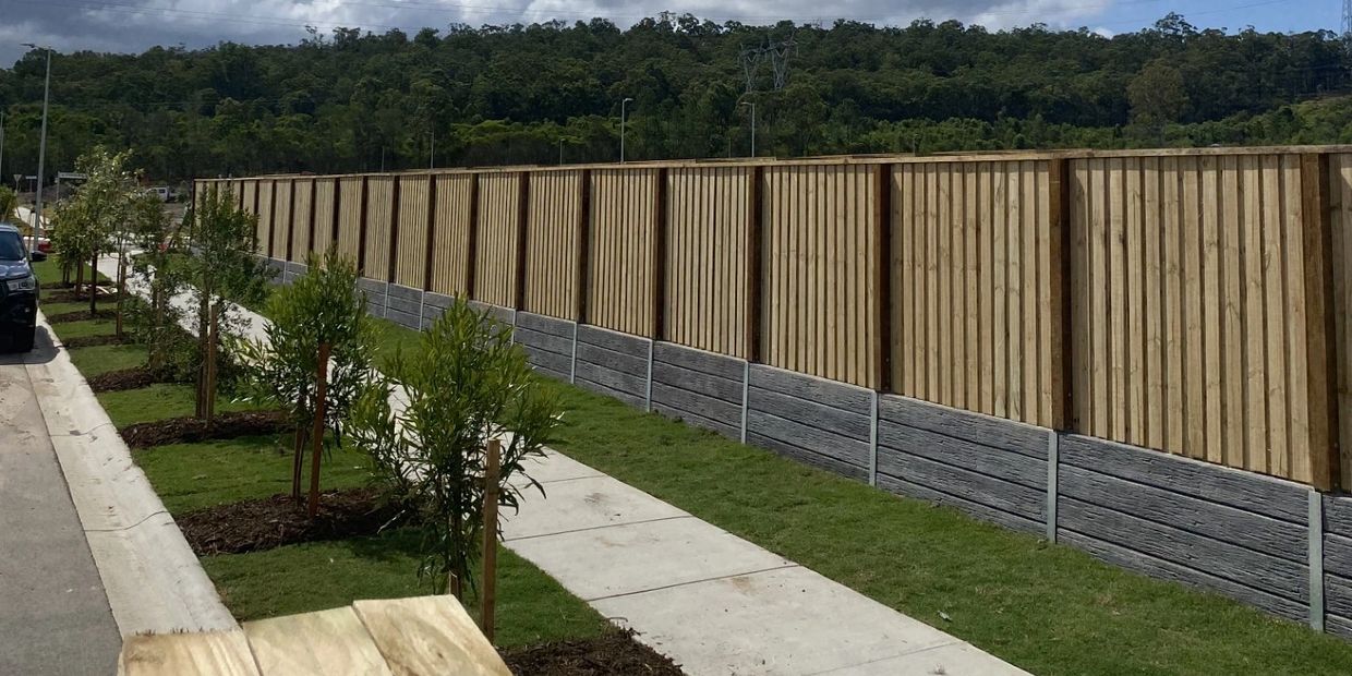 Newly planted trees along a sidewalk beside a wooden fence with a forest backdrop.