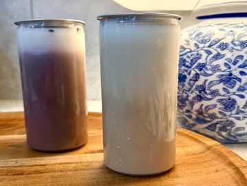Two gradient iced drinks in metal cans on a wooden tray beside a lamp.