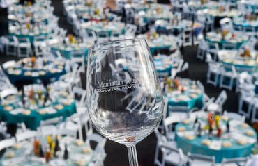A wine glass with 'Manhattan Wine' etched, held over many set tables at an event.