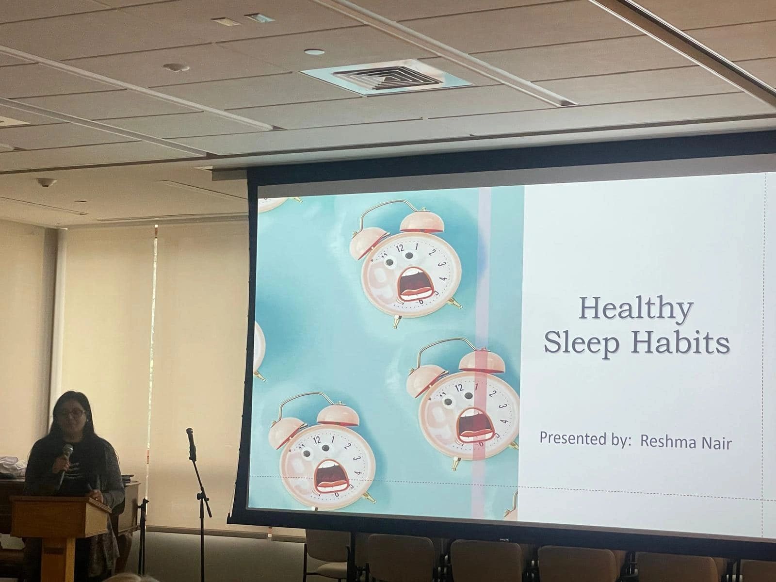 Reshma Air talking in front of screen with Health Sleeo Habits presentation title next to three alarm clock analog faces with mouths opened wide.