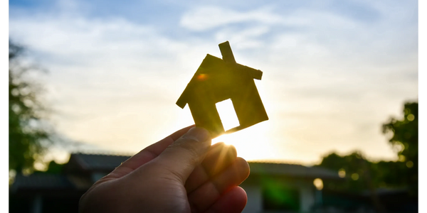 Hand holding a small house cutout against a bright sky at sunset.