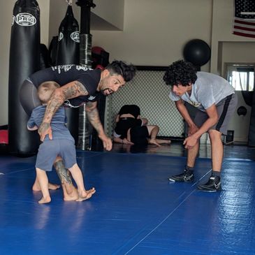 A man teaches wrestling to children in a gym with blue mats and punching bags.