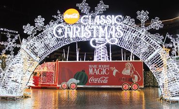 Illuminated Coca-Cola Christmas truck under a festive light arch.