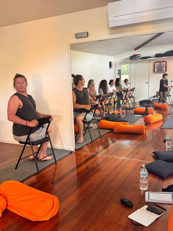 A group yoga class in a mirrored room with orange cushions.