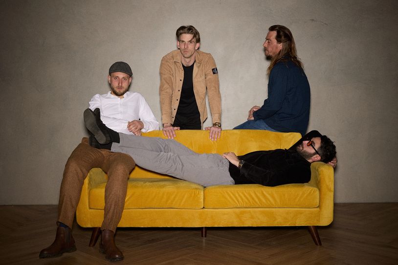 Four men posing with one lying on a yellow sofa and three sitting or standing around it.