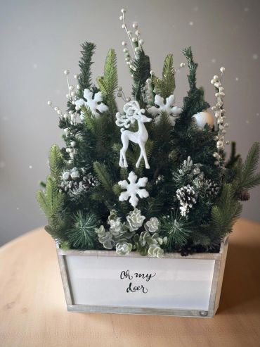 A festive Christmas arrangement with greenery, white snowflakes, and a white reindeer in a planter.