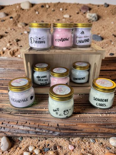 Jars of colorful bath salts with motivational labels on a sandy beach.