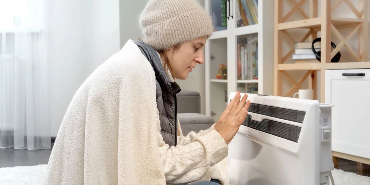 Woman warming her hands by a heater indoors, wrapped in a blanket and wearing a beanie.